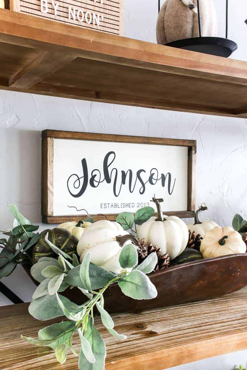 My favorite part of this living room shelf decor is the vintage dough bowl with faux lambs ear greenery, pumpkins, and scented pinecones. Our family sign hangs on the wall behind the bookshelf.