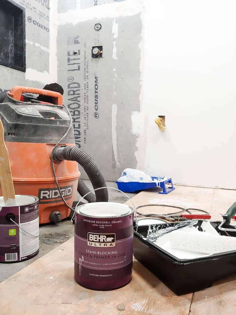 Can of Behr painting sitting on ground next to paint tray with shopvac in the background and concrete backer board up on the walls during a bathroom renovation