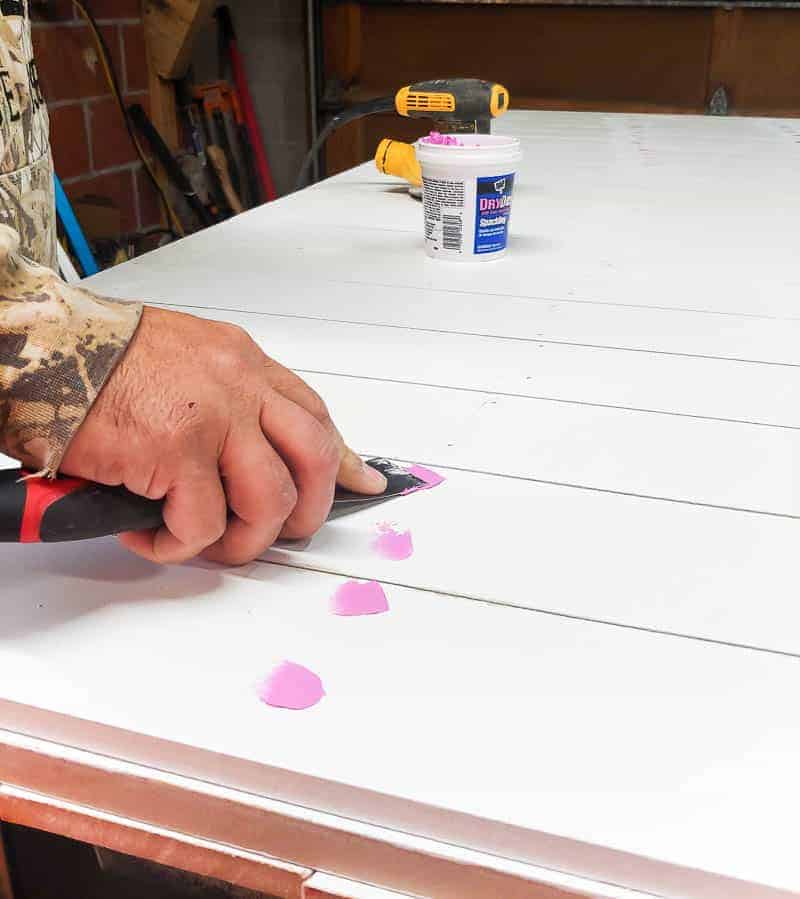 Logan uses a putty knife to fill in the nail holes on the barn door with pink wood filler.