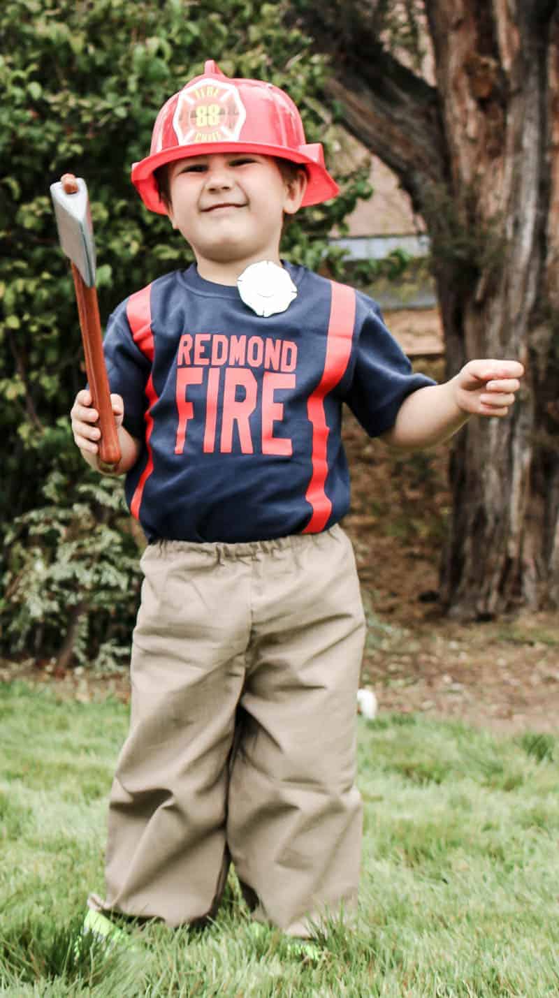 A little boy runs around outside dressed like a firefighter. He's wearing khaki pants with high-vis stripes, a navy blue shirt that says "redmond fire" in red text, and a plastic firefighter helmet. He's holding a toy ax in one hand.