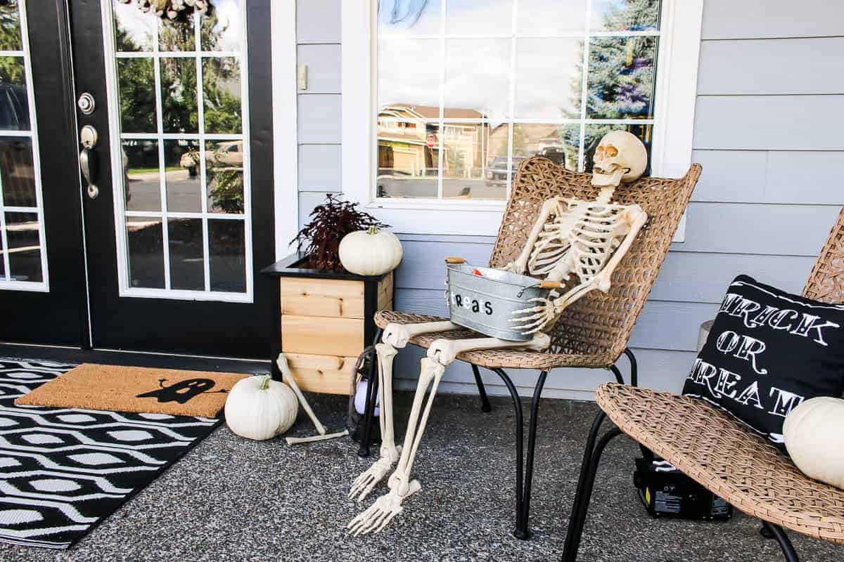 Skeleton holding the metal tub with treats sitting beside another chair with Trick or Treat throw pillow and pumpkins all over for the Halloween Porch Decor