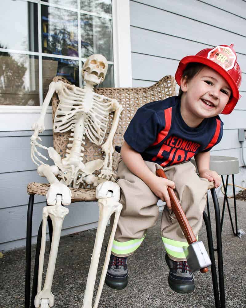 A little toddler boy sits on a chair next to a Halloween skeleton decoration. The boy is dressed up as a firefighter, wearing a shirt, pants, and a plastic red firefighter hat.