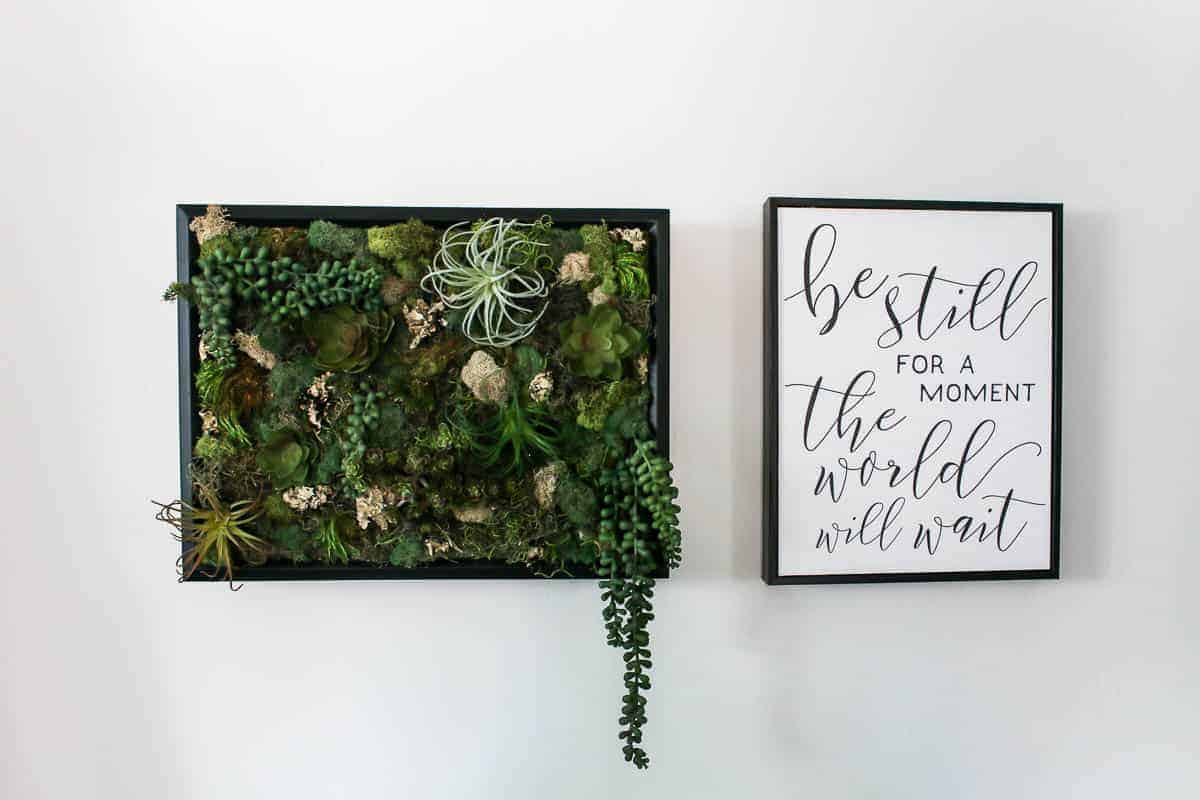 Wall decor shown on white wall with green faux succulents and moss in black frame as a vertical garden and black and white wall decor that says be still for a moment the world will wait