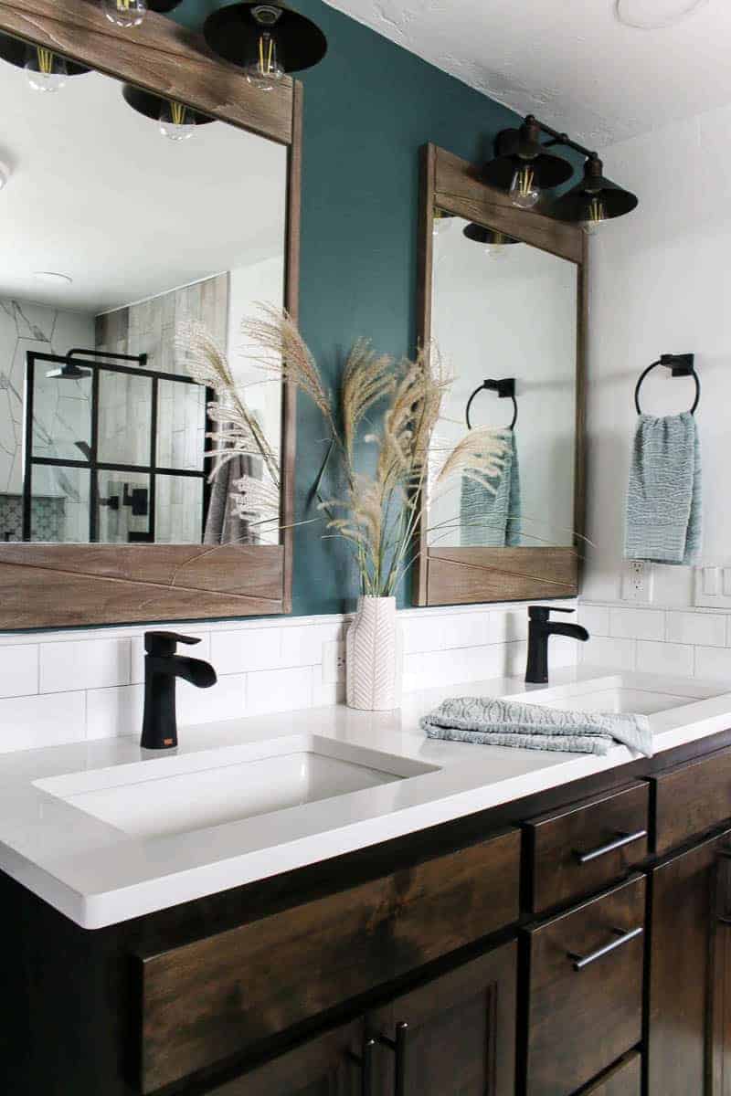 Farmhouse vanity area with dual sinks, black faucets, white quartz countertop and subway tile backsplash, green accent wall, large teak wood mirror and farmhouse style vanity lights