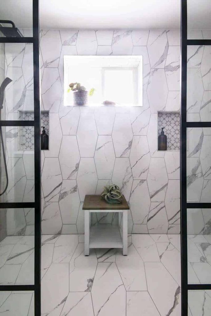Modern looking shower with bright window. White marble tiles and black shower heads with black paneled windows and white table.