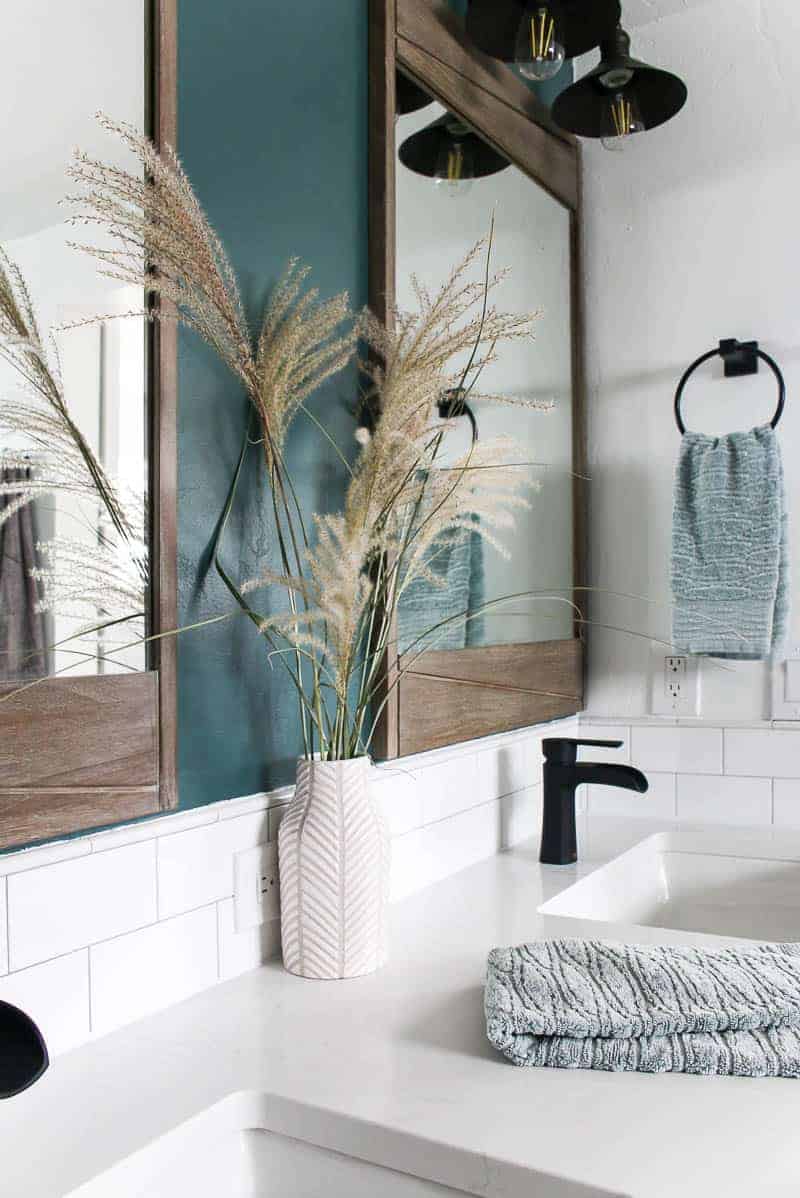 bathroom picture with dried grass in vase and wooden mirror above vanity with black faucets, white quartz countertop with white subway back splash