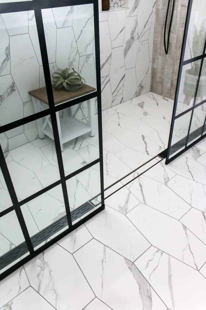 Modern looking shower with white marble tiles and back panel windows and a small table with a plant on it.