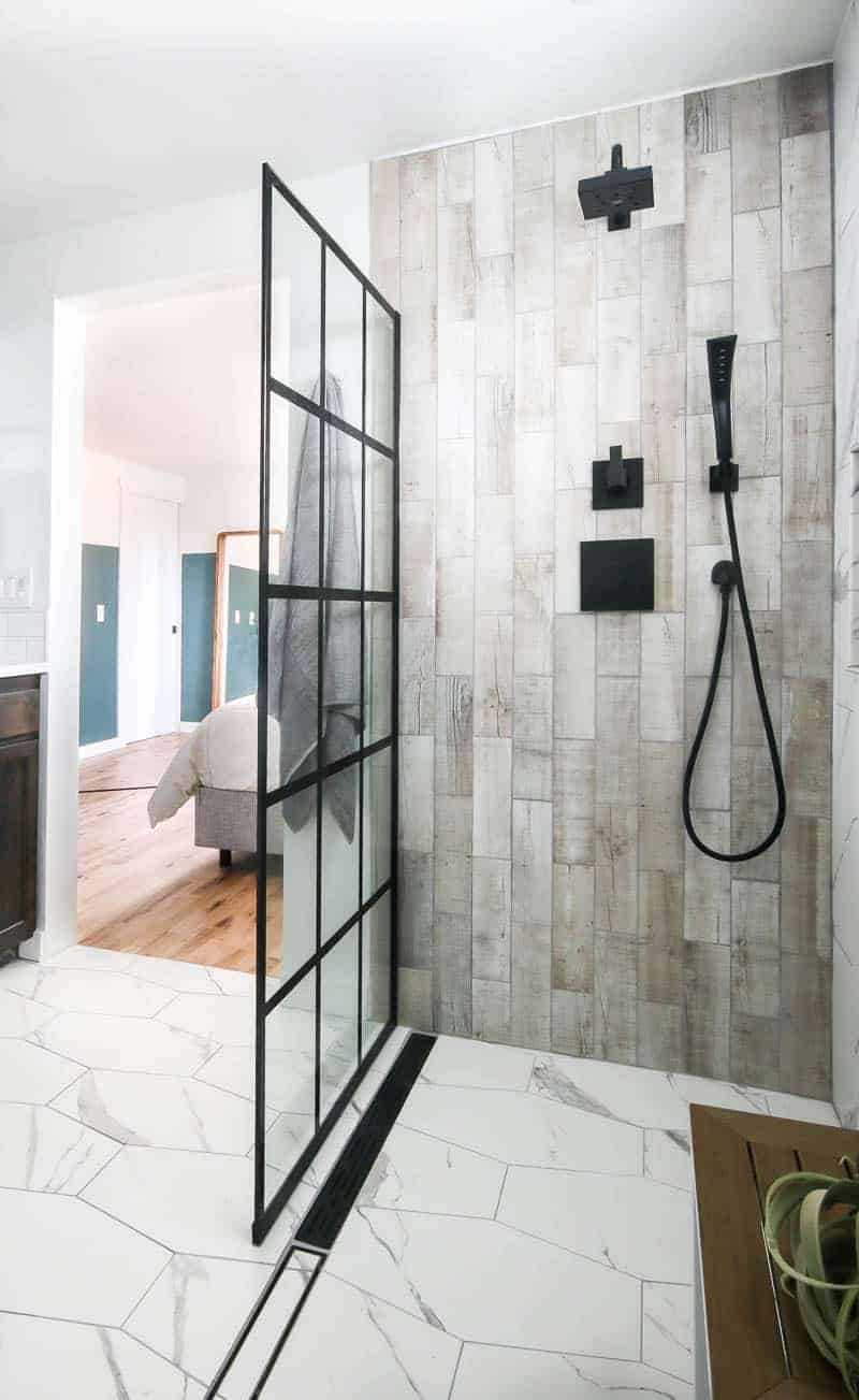 Shower area with wood look tile on sides, black faucet and shower head and white hexagon tile on the floor with windowpane shower door panel. The door way open to the bedroom in the background