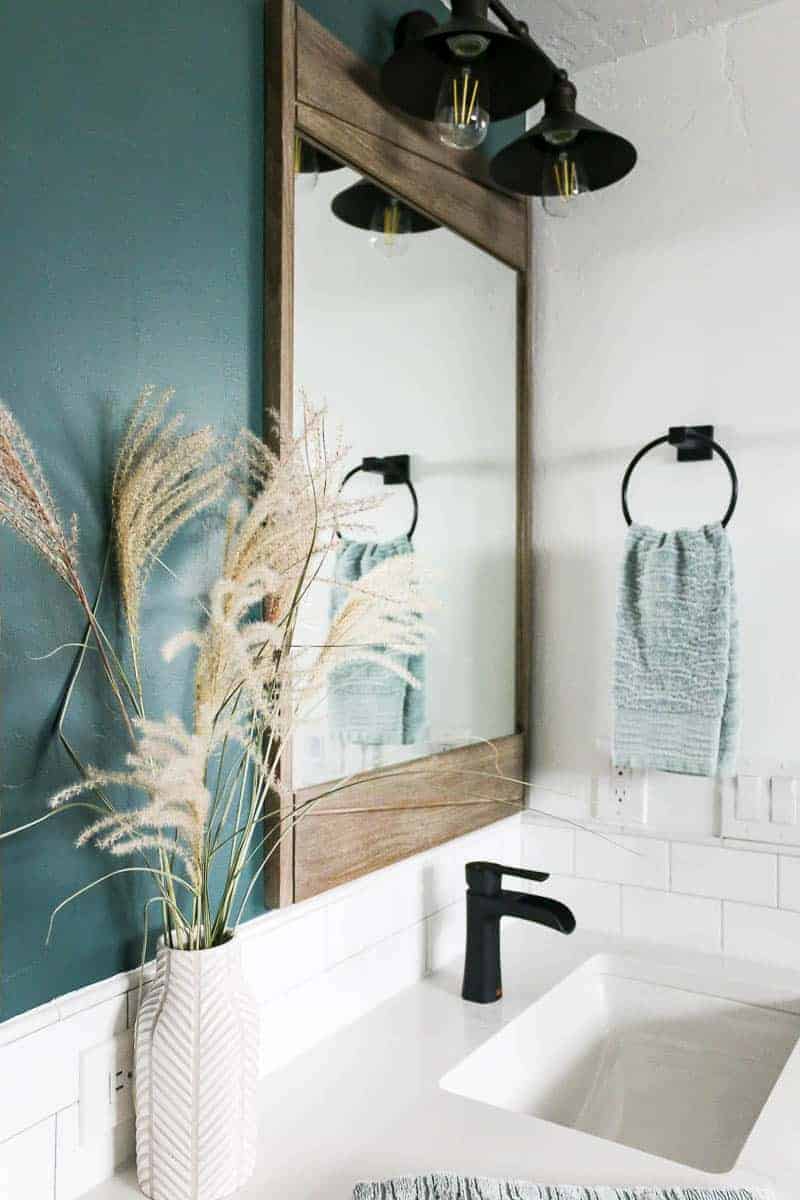 Vanity area in master bathroom with modern farmhouse style showing dark green accent wall, large teak wood mirror, barn vanity light and a towel ring holding green textured hand towel with white sink, countertop, blacksplash and black sink faucet