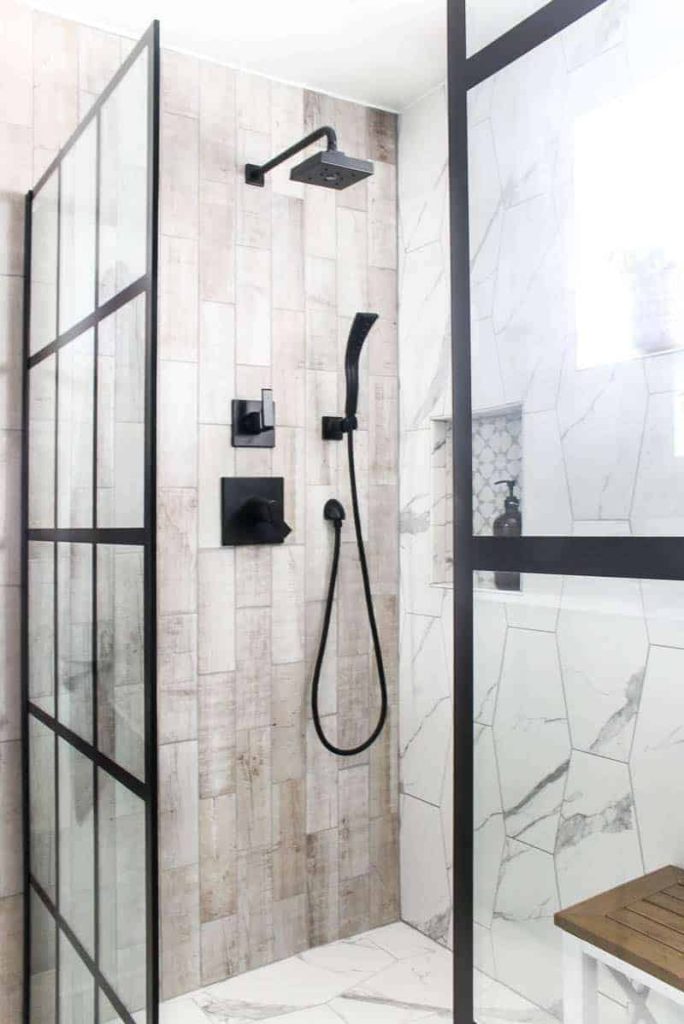 Modern looking shower with white hexagonal marble tile on the right and wood look rectangle tile on the left with a black shower head and windowpane fixed shower door panels.