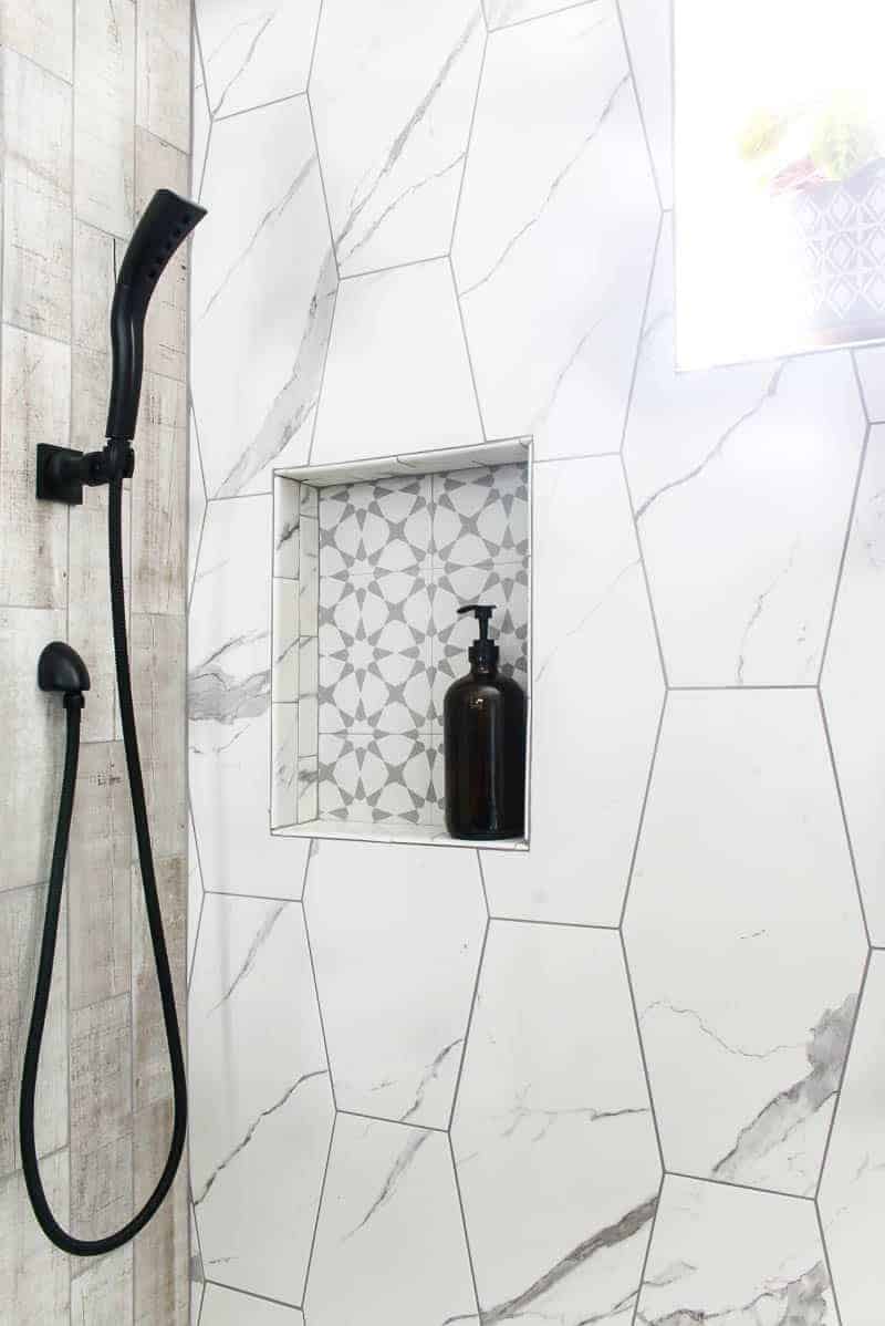 Amber glass soap holder with pump sitting in shower niche in luxurious shower with large white hexagon tile and wood look tile with black hand shower shown on left