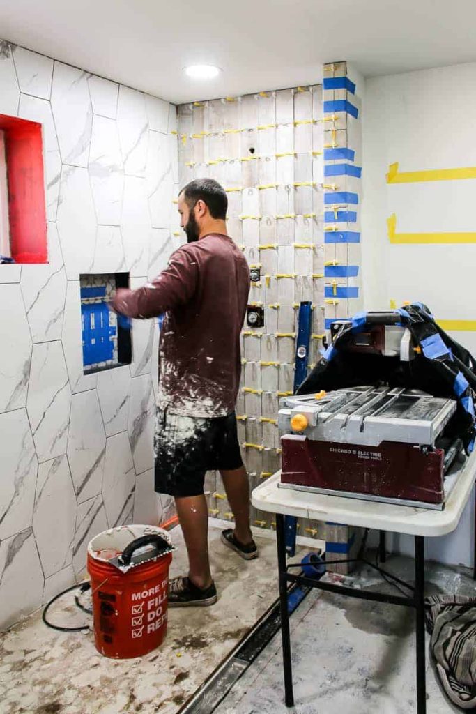Man is putting up hexagonal white marble tile. He is using spacers and blue tape to hold some areas up. There is also a large wet tile saw next to the man. 
