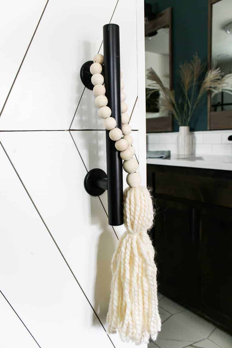 A modern black industrial style door handle with a wooden bead rope accessory, and a bathroom vanity in the background.