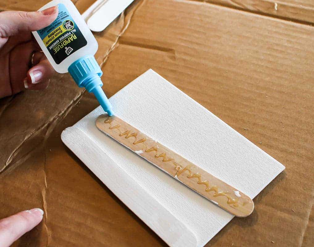 Applying glue at the back of the Popsicle sticks to attach to the canvas.