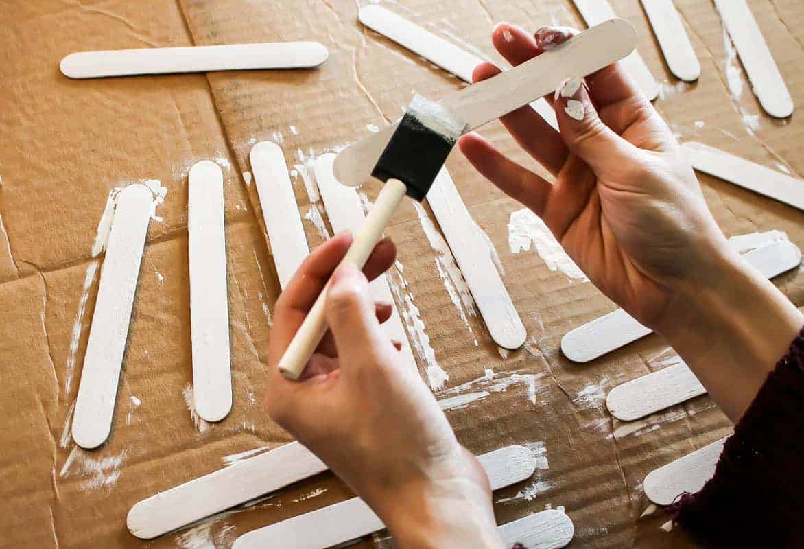 Painting a Popsicle stick with white paint using a foam paint brush and a few more painted Popsicle sticks placed on the carton