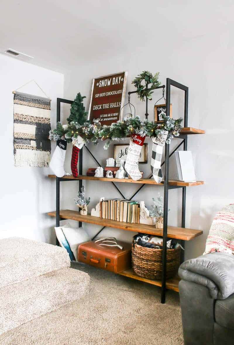 Four layered shelving unit in living room with Snow Day Christmas sign, ornament and farmhouse decorations on the first shelf, on the second shelf are Christmas garlands, Christmas stockings, mini Christamas village with DIY Christmas and personalized family sign, third shelf contains vintage books and corbel bookends and the last shelf has throw pillows, vintage suitcase and a basket with blankets.