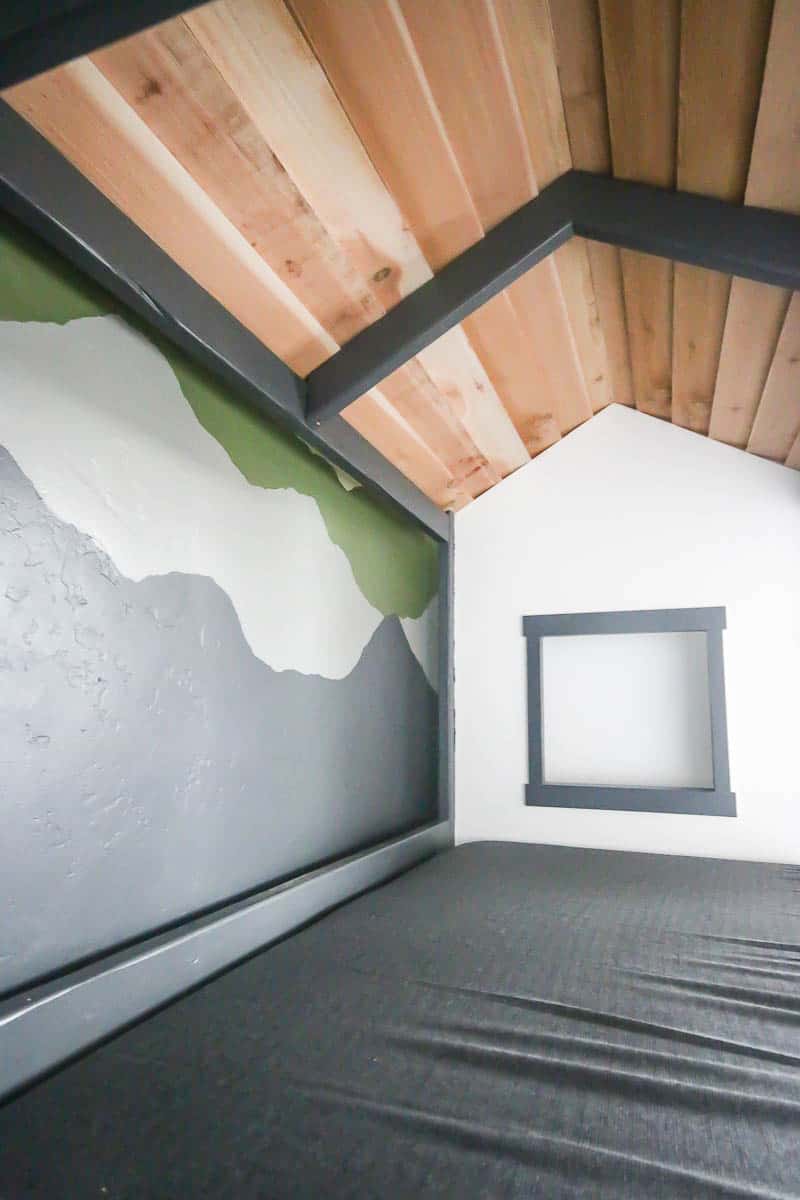 Inside view of the DIY cabin bed showing wooden roof, tiny window at the end and the bed with gray sheets, perfect for a child's first big kid bed