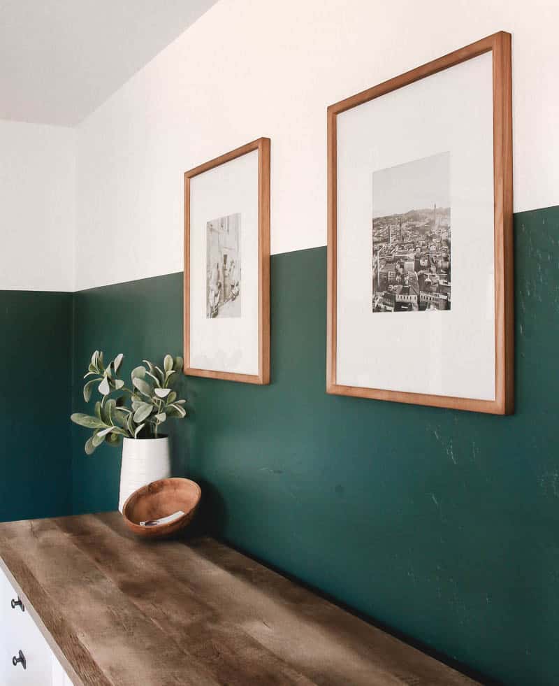 These copper framed landscape photos hang on a two toned white and green bedroom wall, above a wood dresser, On top of the dresser, we have an indoor plant in a sleek white vase and boho style trinkets bowl on a green boho bedroom.
