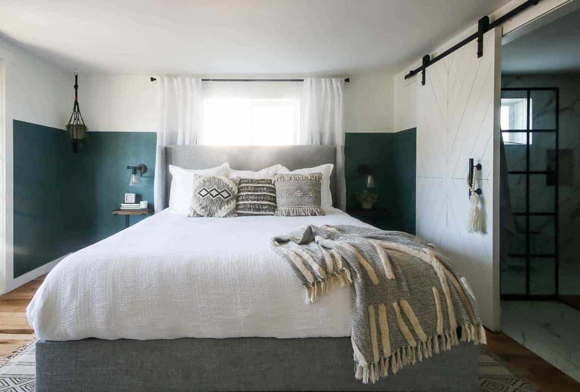A full view of green boho bedroom sits in front of a window, dressed with white linens and boho style throw pillows and throw blanket. To the right, our master bathroom with a DIY sliding barn door.