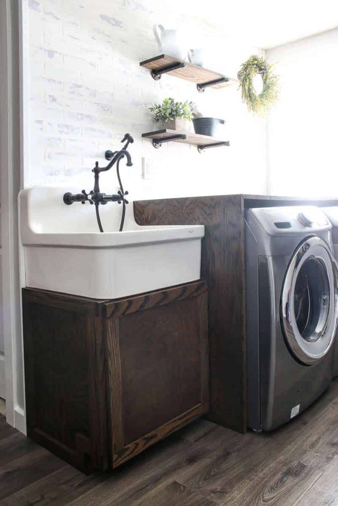 Farmhouse Laundry room with large farmhouse sink sitting on top of a wood cabinet and wood counter covering a washer and dryer with wood floors and white faux brick walls