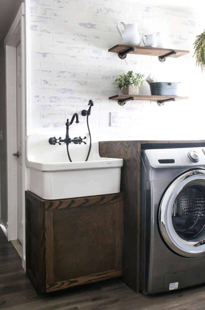 A modern farmhouse laundry room with a farmhouse laundry sink and cabinet, folding table over the washer and dyer, and diy faux brick wall