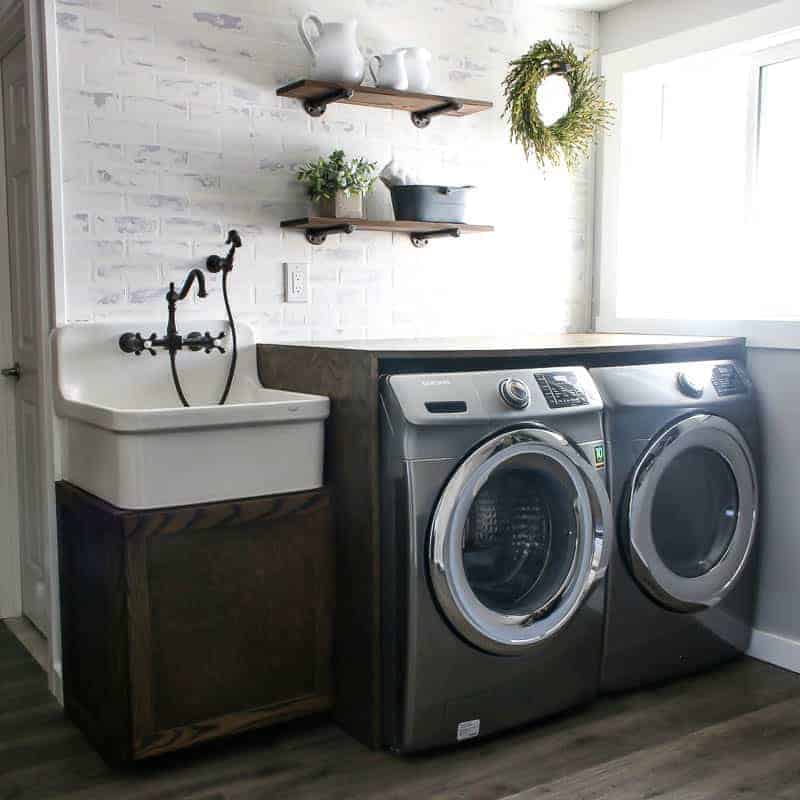 A farmhouse style laundry room with a classic farmhouse sink, industrial shelves on a whitewashed faux brick wall, and washer and dryer with a diy folding table