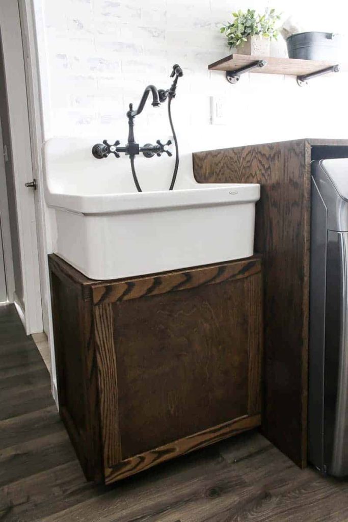 Farmhouse laundry room with large farmhouse sink and vintage faucet and sprayer on top of a wood cabinet with white brink walls and wood floors.