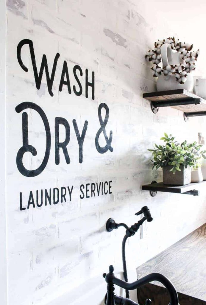 A look at the completed DIY wall stencil, painted on the laundry room wall over the sink, next to farmhouse style shelving with plants and planters.