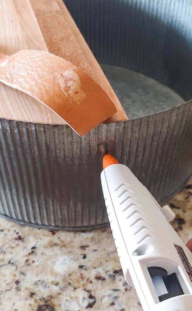 A hot glue gun is gluing a piece of leather to the side of a galvanized basket with a piece of wood at the bottom