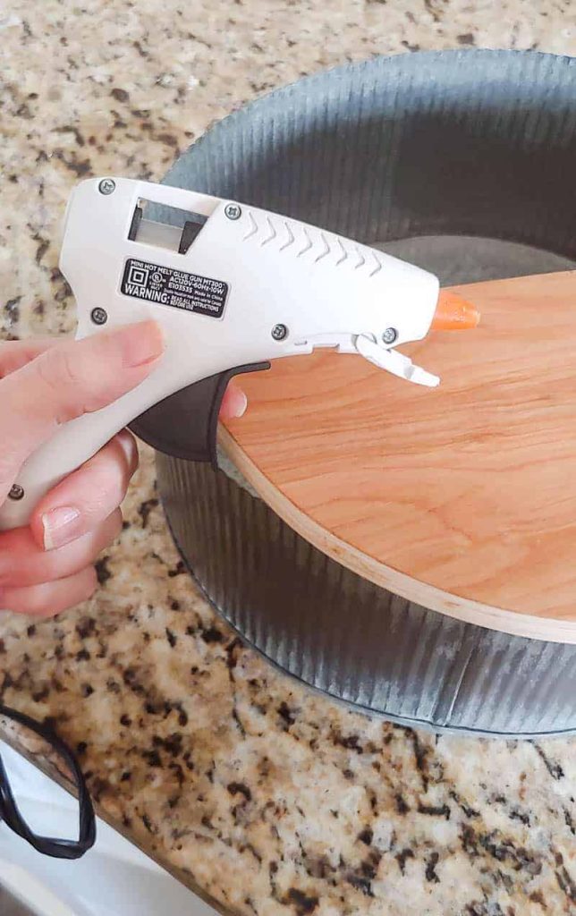 galvanized basket sitting on a counter with a piece of wood in it and a hand holding a glue gun