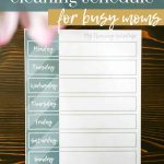shows a weekly cleaning schedule on a wood table with pink flowers in the top corner with overlay texts that says free printable cleaning schedule for busy moms, download here