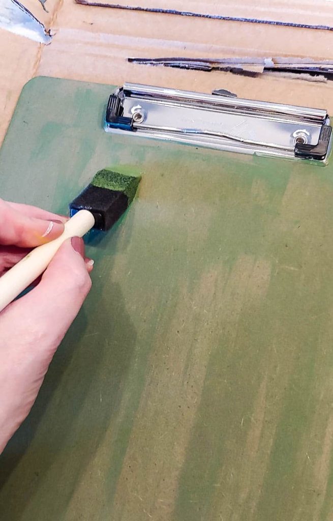 Shows a hand painting a clipboard on cardboard with green paint