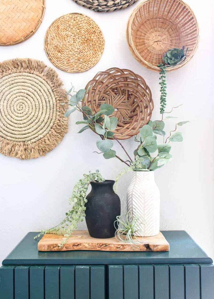 My upcycled black vintage vase and white ceramic vase hold lambs ear and eucalyptus leaves, and sit under a wicker basket wall in my living room. 