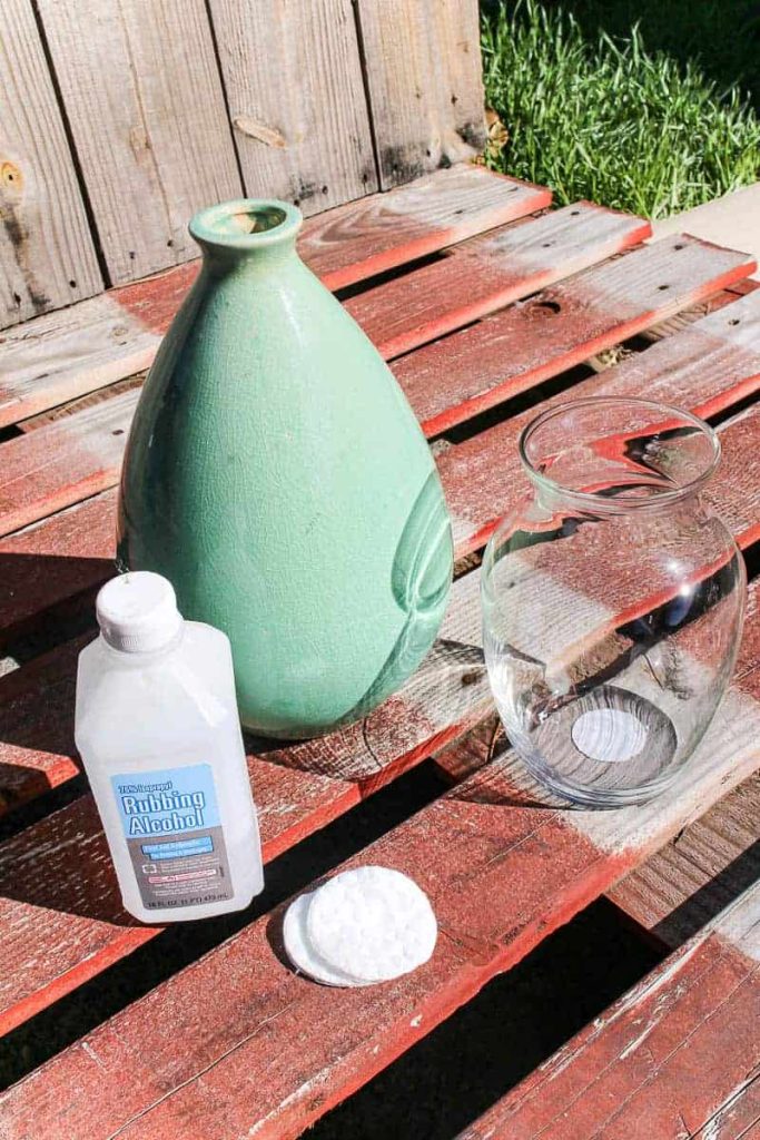 Two vases sit outside on a picnic table next to a bottle of rubbing alcohol and a cotton swab. Use the alcohol to clean the vases so the paint easily adheres.