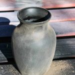 shows a black vase with dirt on it outside on a wood pallet