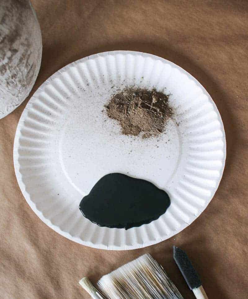 A paper plate with a pile of dirt and a pool of black craft paint are the supplies needed for the DIY aged vase