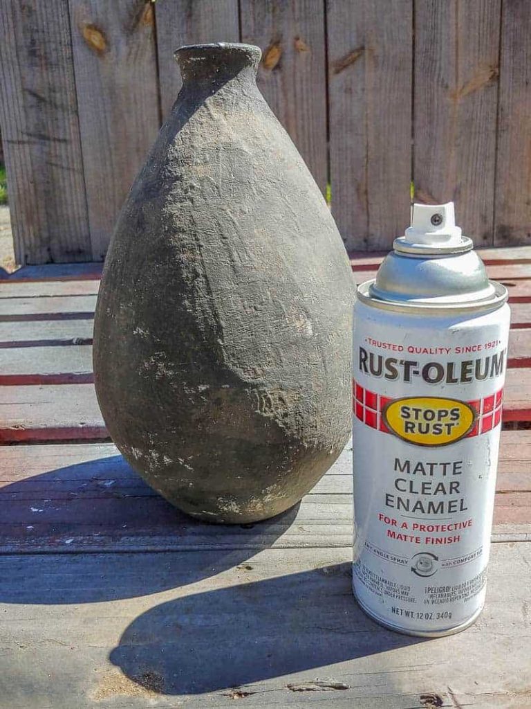 A black and brown vase sits on a wooden table, and a can of matte clear enamel finish sitting next to the upcycled vase.