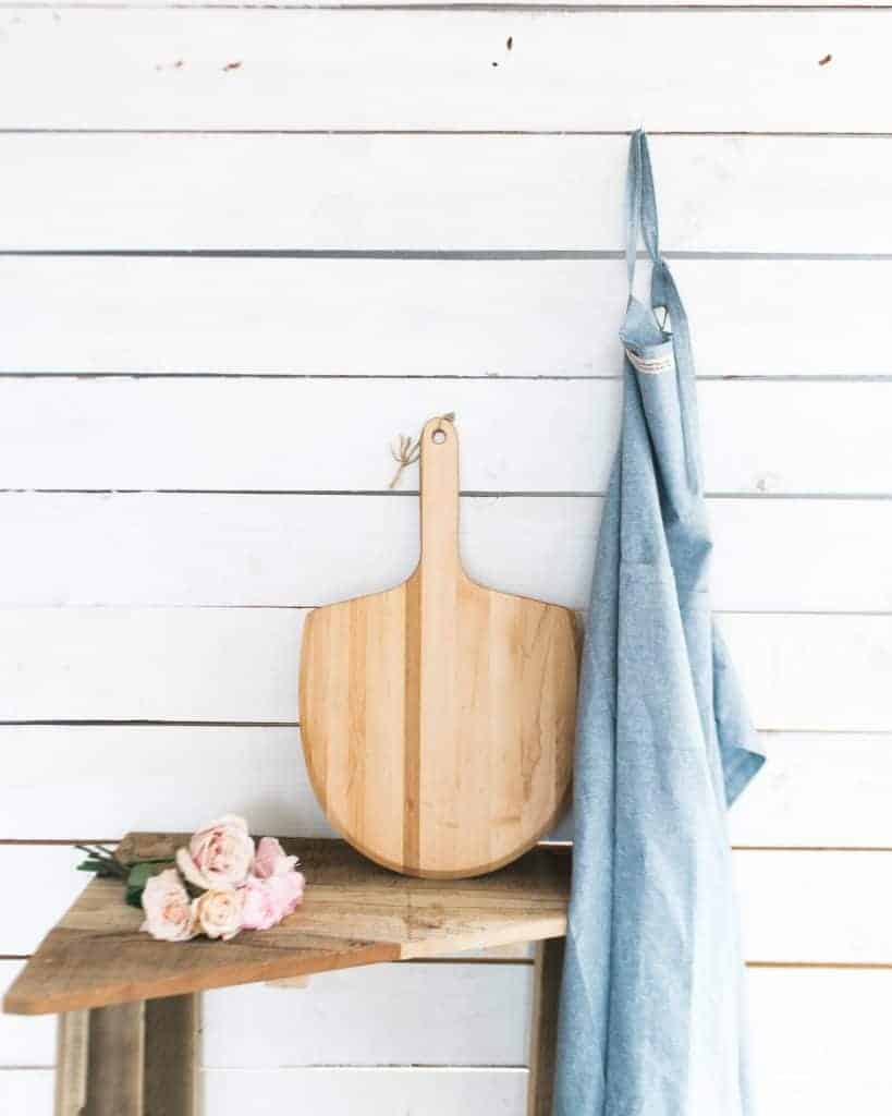 rustic shiplap wall painted white with wooden stool and wooden cutting board and roses on it with a blue apron hanging on the wall