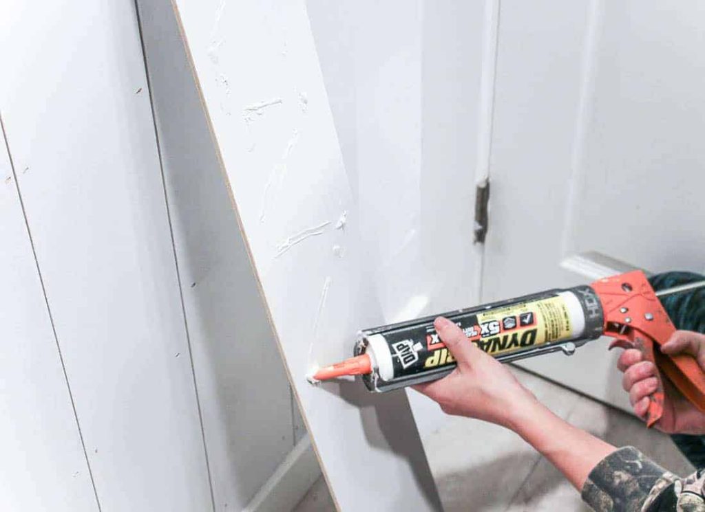 Using Dynagrip construction adhesive to adhere the slats of white painted vertical shiplap to the bathroom wall.