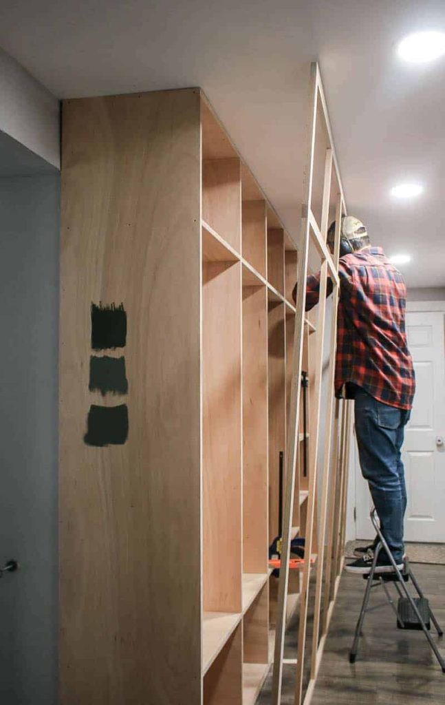 shows a man holding up a face frame while attaching to build in mudroom cabinets on a step ladder in a hallway with gray wood floors