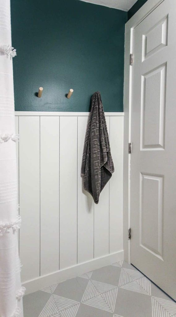 The completed vertical shiplap accent wall in our bathroom; half the wall is shiplap painted white, and the top half is painted a deep teal. Three towel pegs hang on the wall, one holding a bath towel.