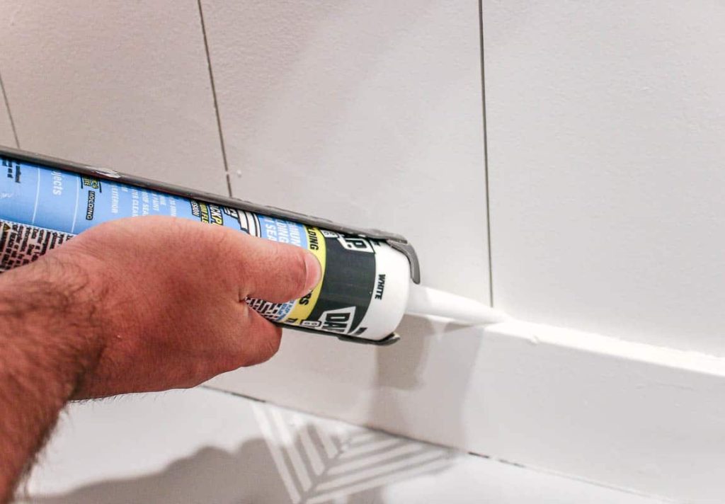 Using caulk to seal the baseboards on the white vertical shiplap wall.