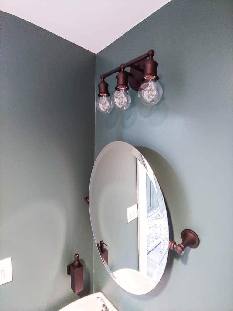 shows a side angled view a dark teal bathroom with an oval mirror and farmhouse styled vanity lights