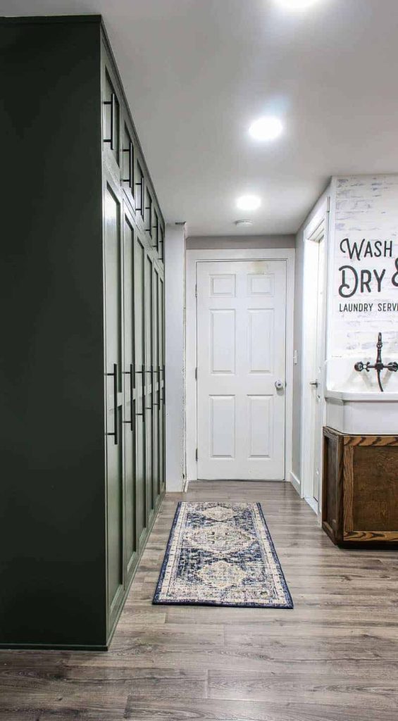 shows a farmhouse green mudroom lockers with blue and white boho runner rug on the gray wood floors in a white hallway and door with wash and dry laundry service stenciled on the wall