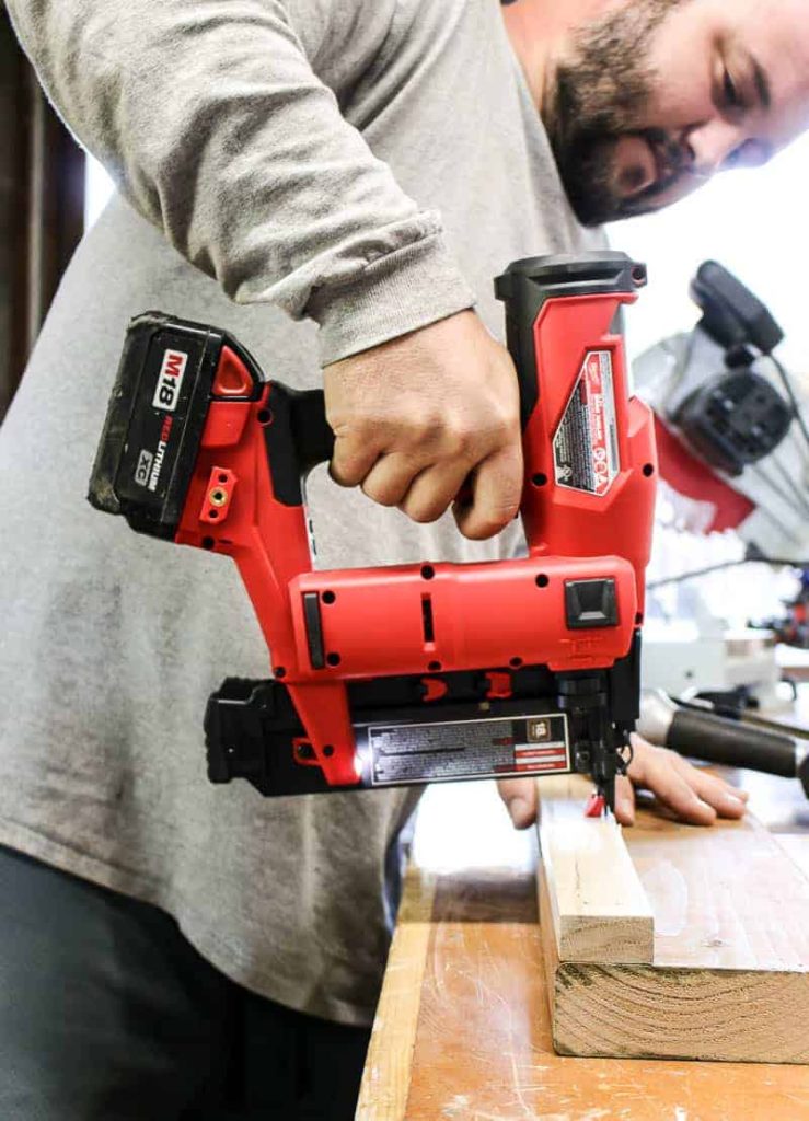 shows a man in a gray long sleeve using a cordless Milwaukee m18 nailer on a piece of wood on a table during a DIY woodworking project.