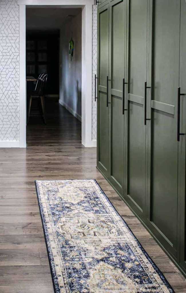 shows a farmhouse green mudroom lockers with blue and white boho runner rug on the gray wood floors in a white wallpapered wall and kitchen in background