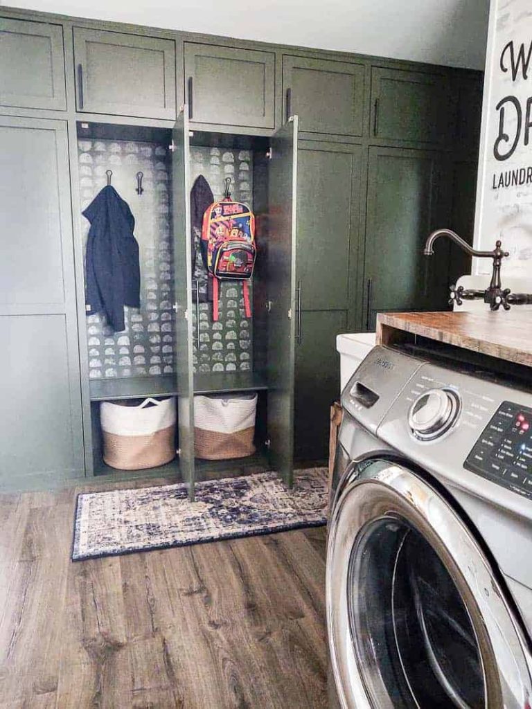 shows a farmhouse laundry room with green farmhouse mudroom lockers with some jackets and backpacks with white sponge painted spots in the back with a boho runner rug on the gray wood floors