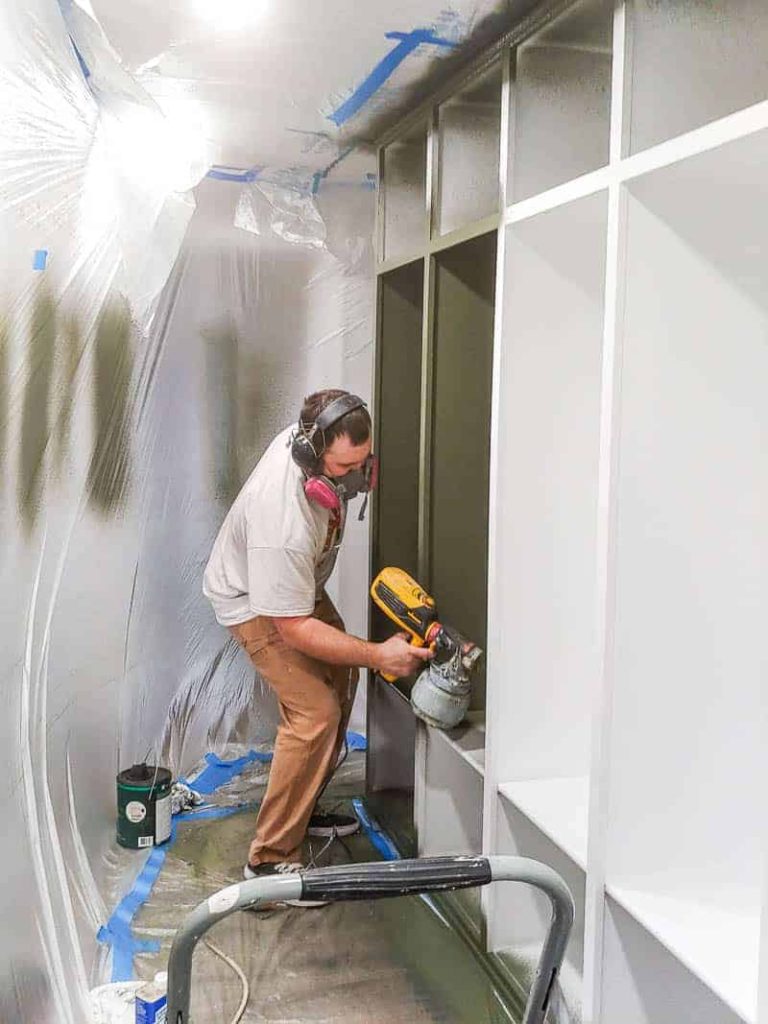 shows a man using a paint sprayer to paint the shelves of the mudroom green with the rest of the room covered in plastics and tape