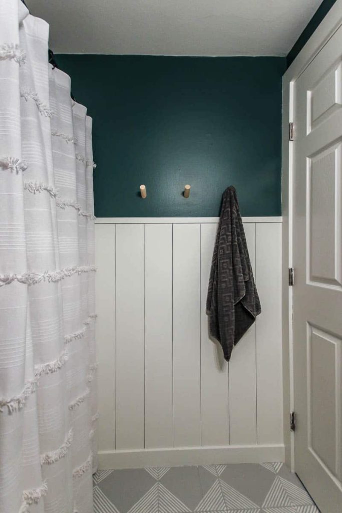 The accent wall in our bathroom; half of the wall has white painted vertical shiplap, and the top half is painted teal with towel holders. A white linen show curtain hangs on the left side.