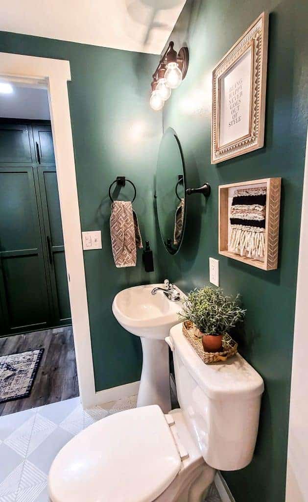 A look from the inside of our new Buget Bathroom Makeover shows the vanity, freestanding sink, round glass mirror, and boho style wall art hanging on deep green painted bathroom walls.